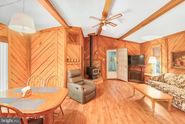 living room with light wood-type flooring, lofted ceiling with beams, a wood stove, ceiling fan, and wooden walls