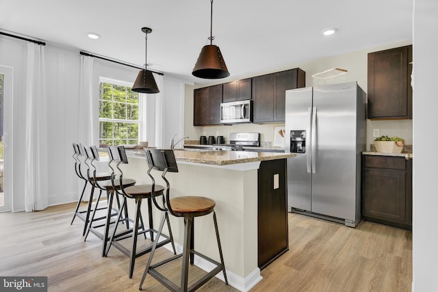 kitchen with dark brown cabinets, light wood-type flooring, hanging light fixtures, a center island with sink, and appliances with stainless steel finishes