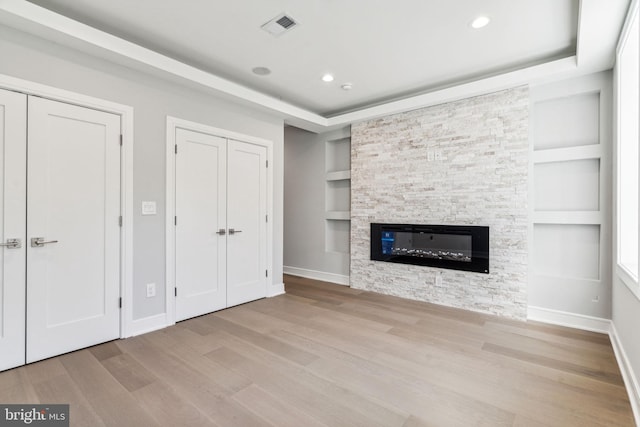 unfurnished living room featuring a stone fireplace, light hardwood / wood-style flooring, a tray ceiling, and built in shelves