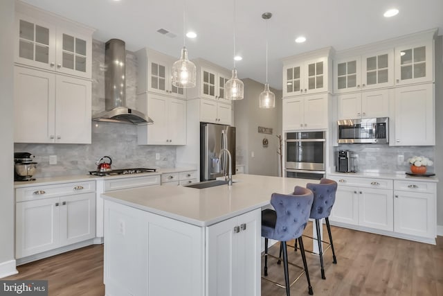 kitchen with pendant lighting, wall chimney exhaust hood, a kitchen island with sink, white cabinetry, and appliances with stainless steel finishes