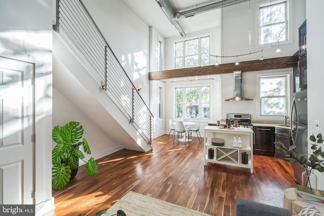 kitchen with a high ceiling, wall chimney range hood, dark wood-type flooring, a healthy amount of sunlight, and stainless steel gas range