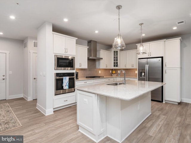 kitchen with an island with sink, sink, wall chimney exhaust hood, white cabinetry, and stainless steel appliances