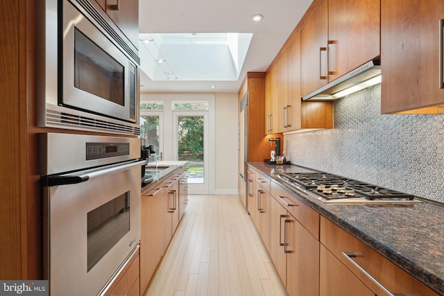 kitchen featuring a skylight, stainless steel appliances, dark stone countertops, light wood-type flooring, and decorative backsplash