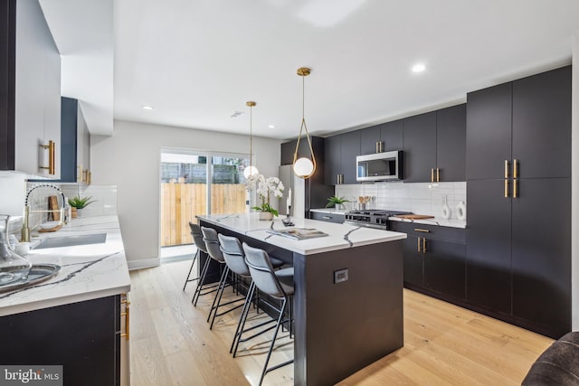 kitchen with an island with sink, hanging light fixtures, backsplash, appliances with stainless steel finishes, and light wood-type flooring