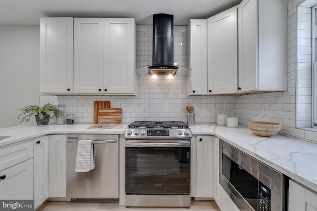 kitchen featuring white cabinets, wall chimney range hood, decorative backsplash, appliances with stainless steel finishes, and light stone counters