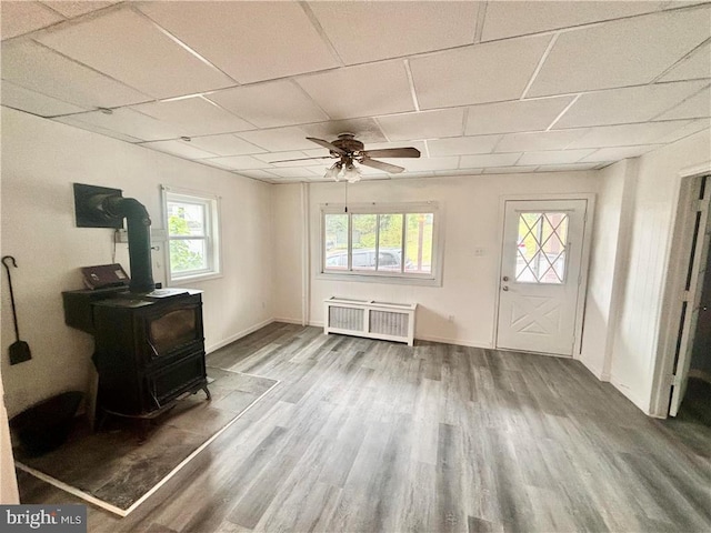 interior space with a drop ceiling, a wood stove, ceiling fan, and hardwood / wood-style floors