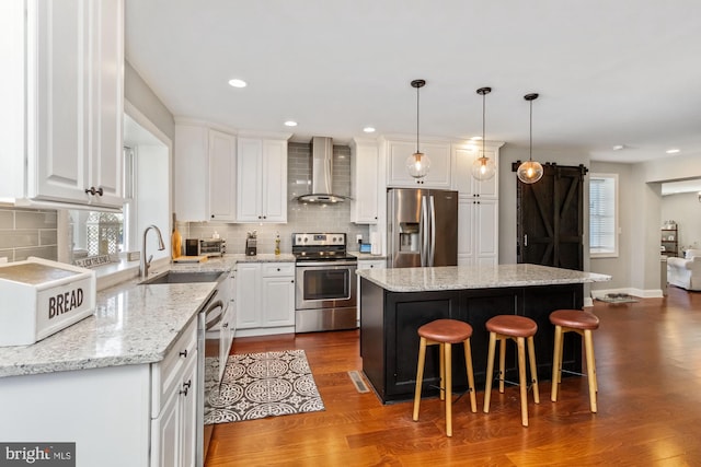 kitchen with wood-type flooring, appliances with stainless steel finishes, a barn door, a center island, and wall chimney exhaust hood