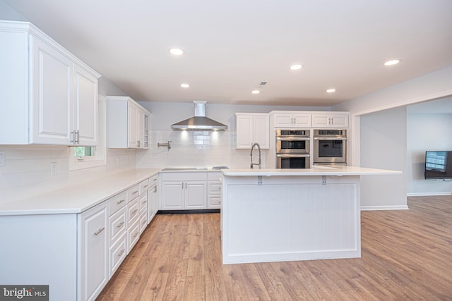 kitchen featuring stainless steel double oven, light hardwood / wood-style floors, wall chimney range hood, and white cabinetry