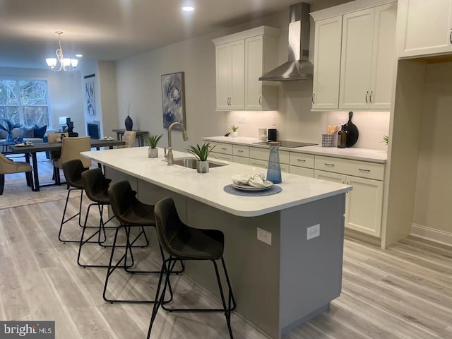 kitchen featuring wall chimney range hood, sink, light wood-type flooring, and a center island with sink