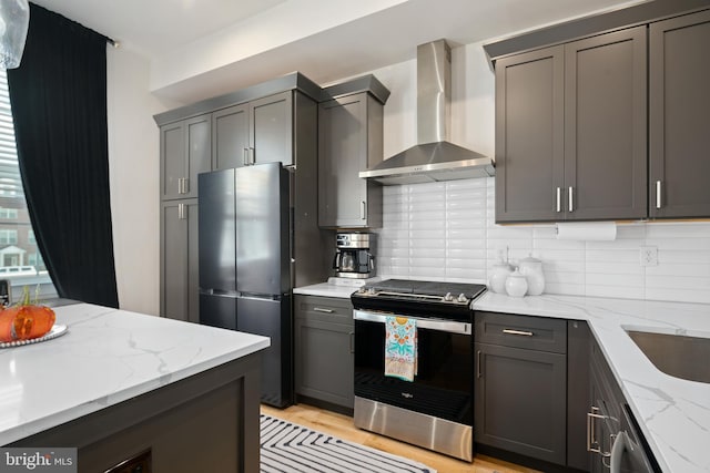 kitchen with wall chimney range hood, stainless steel appliances, light stone countertops, light hardwood / wood-style floors, and decorative backsplash