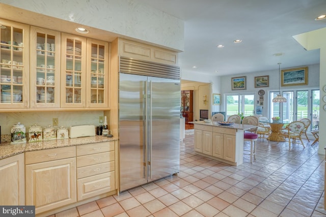 kitchen with light brown cabinetry, light stone countertops, stainless steel built in refrigerator, and decorative light fixtures