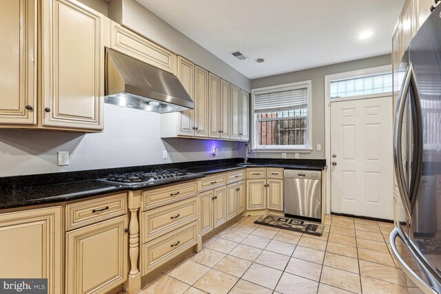 kitchen featuring light tile patterned floors, dark stone countertops, wall chimney range hood, and stainless steel appliances