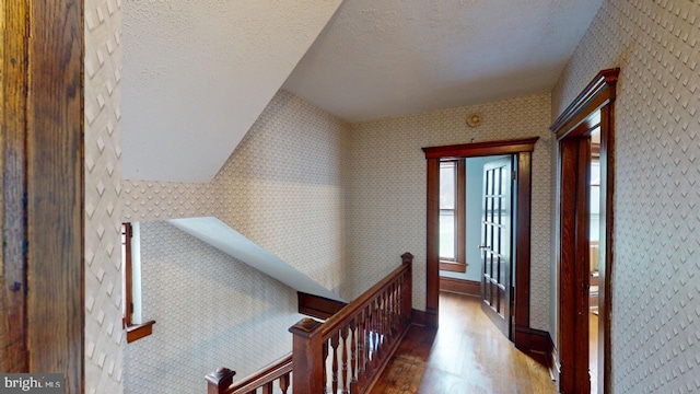 hallway with a textured ceiling, baseboards, wood finished floors, and an upstairs landing
