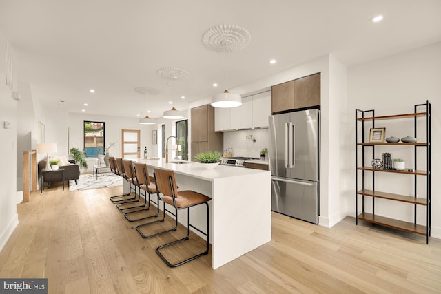 kitchen with sink, high end refrigerator, pendant lighting, a kitchen island with sink, and light wood-type flooring