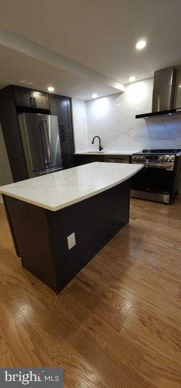 kitchen featuring appliances with stainless steel finishes, decorative backsplash, light stone countertops, light wood-type flooring, and wall chimney range hood