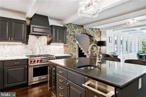 kitchen with high end stainless steel range, beamed ceiling, a center island with sink, and wall chimney exhaust hood