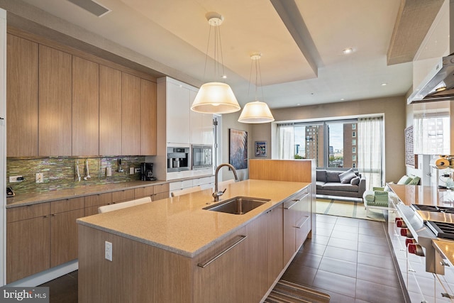 kitchen featuring dark tile patterned flooring, an island with sink, hanging light fixtures, backsplash, and sink