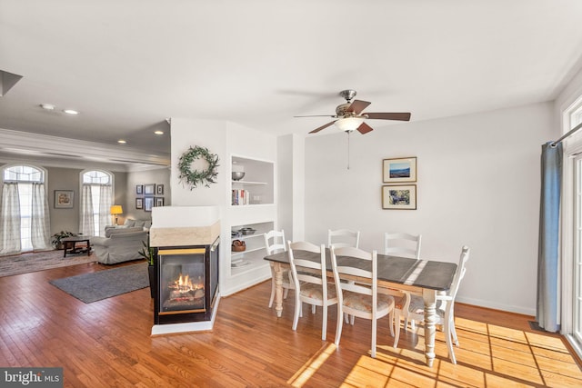 dining room with light wood-type flooring, built in features, baseboards, a multi sided fireplace, and ceiling fan