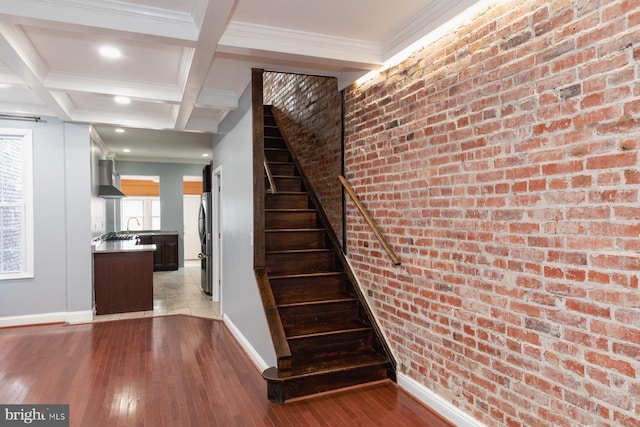 stairway with beamed ceiling, wood-type flooring, brick wall, crown molding, and coffered ceiling