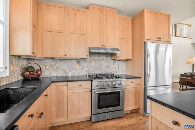kitchen with light wood finished floors, light brown cabinetry, dark countertops, under cabinet range hood, and appliances with stainless steel finishes