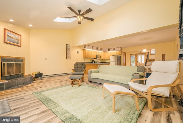 living room with ceiling fan, a skylight, a baseboard heating unit, a tile fireplace, and light wood-type flooring