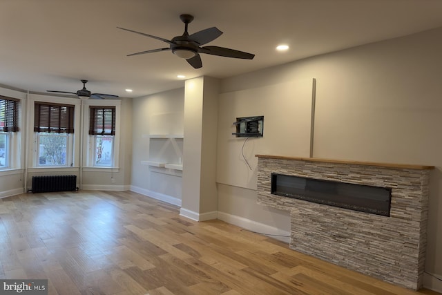 unfurnished living room featuring ceiling fan, a stone fireplace, radiator, and light hardwood / wood-style flooring