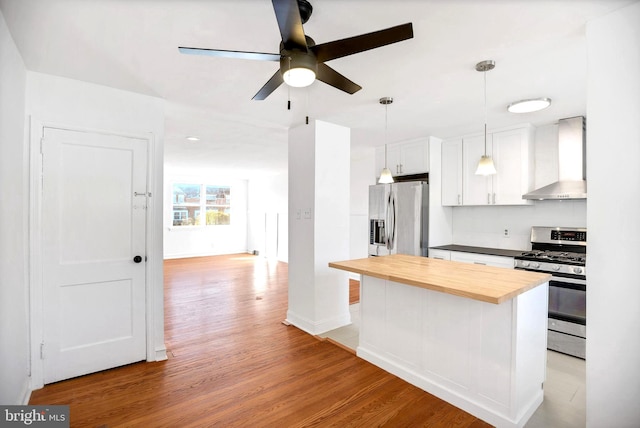kitchen with wall chimney range hood, light wood-style flooring, appliances with stainless steel finishes, ceiling fan, and wooden counters