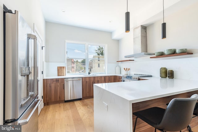 kitchen featuring hanging light fixtures, kitchen peninsula, wall chimney range hood, appliances with stainless steel finishes, and light hardwood / wood-style floors