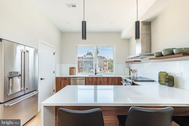 kitchen with wall chimney exhaust hood, pendant lighting, kitchen peninsula, appliances with stainless steel finishes, and a kitchen breakfast bar