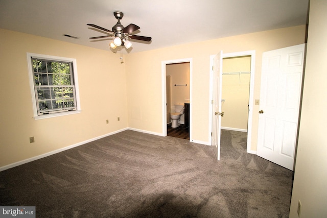 unfurnished bedroom with ensuite bath, a walk in closet, ceiling fan, and dark carpet