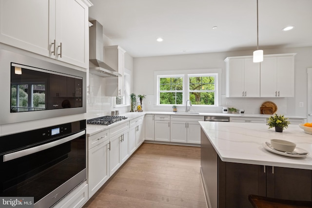 kitchen featuring wall chimney range hood, sink, hanging light fixtures, white cabinetry, and stainless steel appliances