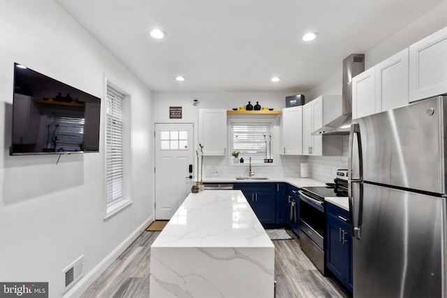 kitchen with stainless steel appliances, sink, blue cabinetry, wall chimney range hood, and white cabinetry