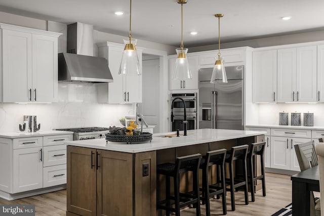 kitchen featuring wall chimney range hood, appliances with stainless steel finishes, white cabinetry, and a kitchen island with sink