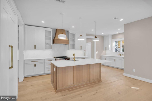 kitchen with premium range hood, a kitchen island with sink, sink, decorative light fixtures, and white cabinets