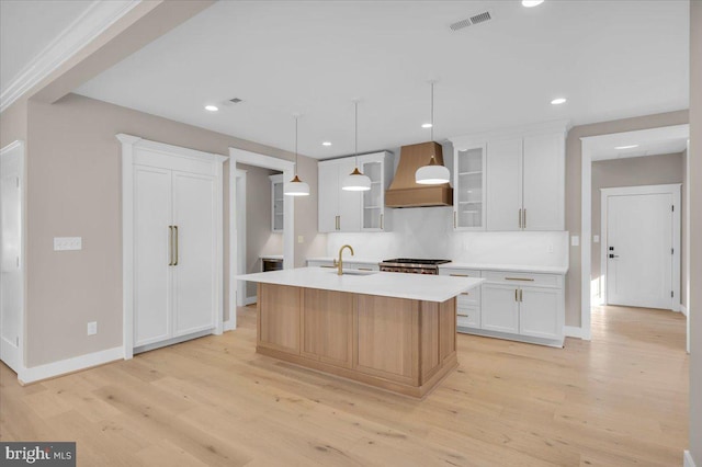 kitchen featuring light wood-type flooring, premium range hood, a center island with sink, white cabinetry, and hanging light fixtures