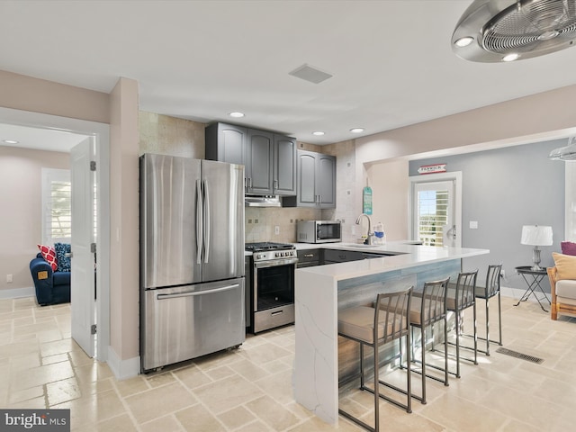 kitchen featuring gray cabinets, tasteful backsplash, kitchen peninsula, appliances with stainless steel finishes, and a kitchen bar