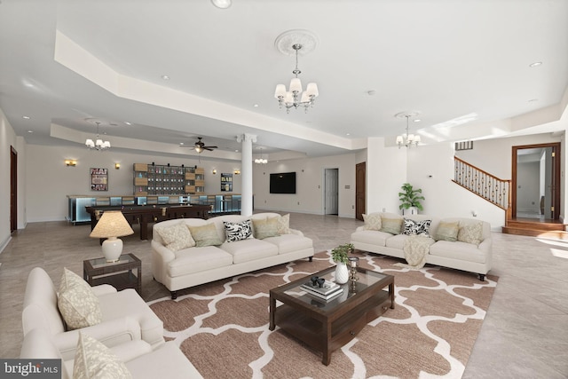 living room with baseboards, a raised ceiling, stairs, ornate columns, and ceiling fan with notable chandelier