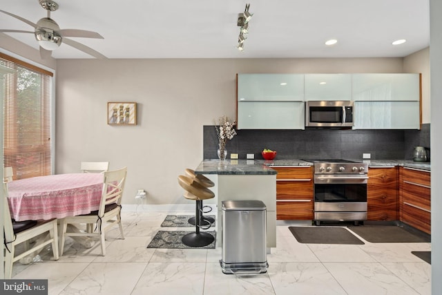 kitchen featuring appliances with stainless steel finishes, dark stone counters, ceiling fan, and backsplash