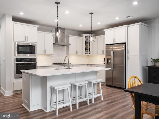 kitchen with a kitchen island with sink, appliances with stainless steel finishes, wall chimney exhaust hood, and white cabinetry