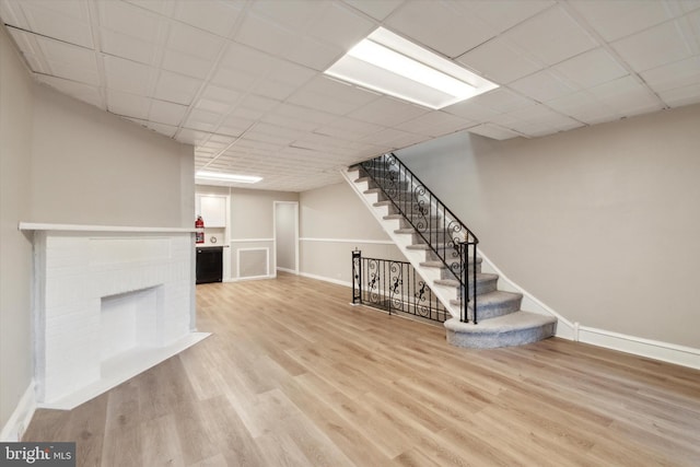 basement with a drop ceiling, a brick fireplace, and light hardwood / wood-style flooring