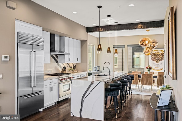 kitchen featuring an island with sink, beamed ceiling, high end appliances, and wall chimney exhaust hood