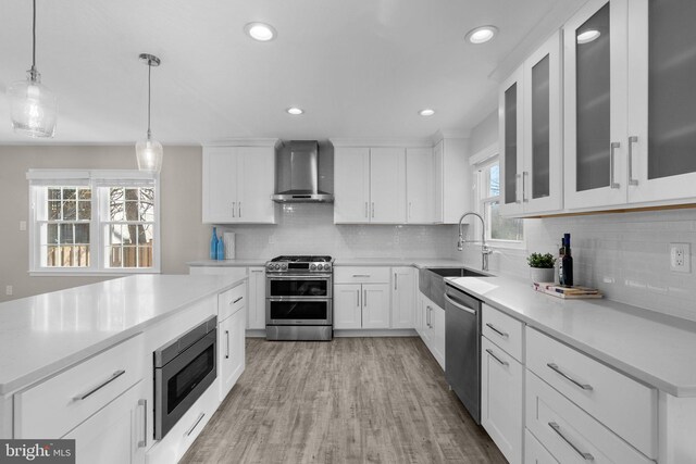 kitchen featuring appliances with stainless steel finishes, wall chimney exhaust hood, sink, white cabinetry, and decorative light fixtures