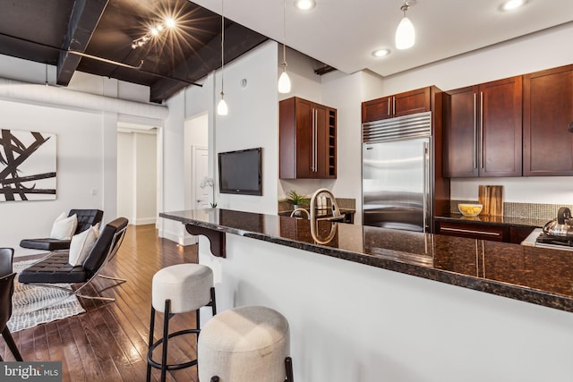 kitchen with pendant lighting, dark wood-type flooring, built in fridge, dark stone countertops, and a kitchen bar