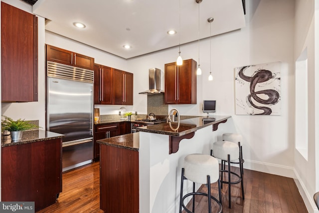 kitchen featuring kitchen peninsula, appliances with stainless steel finishes, wall chimney exhaust hood, dark wood-type flooring, and pendant lighting