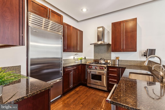 kitchen with dark stone counters, wall chimney exhaust hood, high end appliances, dark wood-type flooring, and sink