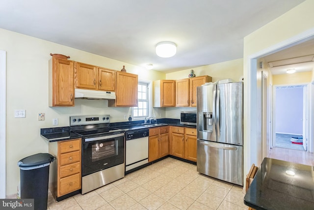 kitchen featuring a sink, light tile patterned flooring, dark countertops, under cabinet range hood, and appliances with stainless steel finishes