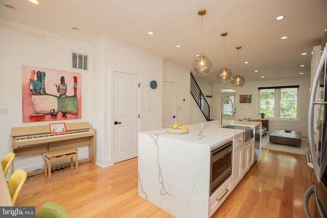 kitchen featuring light stone counters, ornamental molding, decorative light fixtures, a center island with sink, and light hardwood / wood-style floors