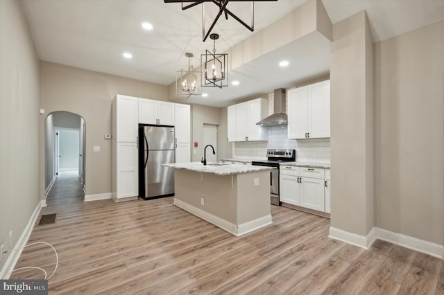 kitchen with stainless steel appliances, wall chimney exhaust hood, light hardwood / wood-style floors, white cabinetry, and a center island with sink