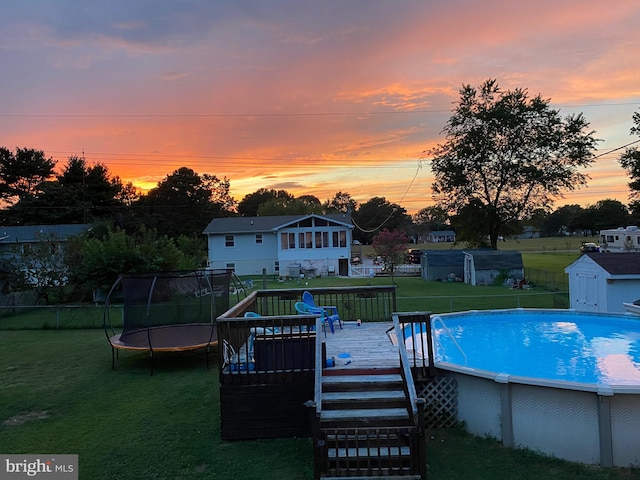 pool at dusk featuring a deck, a trampoline, a yard, and a shed