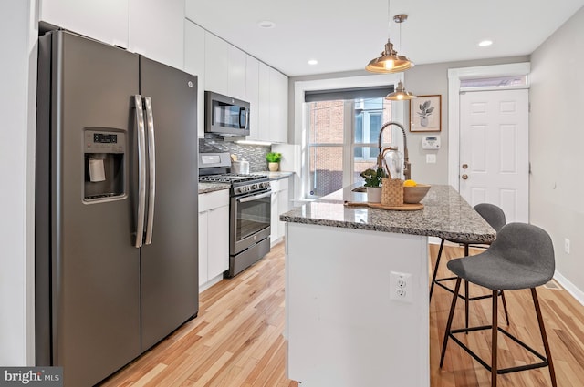 kitchen with an island with sink, light wood-type flooring, hanging light fixtures, white cabinetry, and appliances with stainless steel finishes